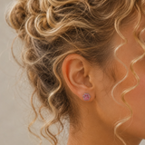 A close-up side profile of a girl with curly blonde hair wearing a small pink paw-shaped stud earring with a white outline and tiny light-colored details.