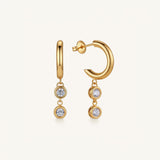A pair of gold huggie hoop earrings with dangling triple-stone drops, shown on a clean off-white background.