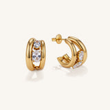 A pair of gold huggie hoop earrings with clear gemstones, shown on a clean off-white background. One earring is facing forward, displaying a central cluster of three round-cut clear stones, while the other is turned to show the side profile with a marquise-cut clear stone and a wing-like gold accent.