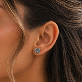 A close-up of a girl's ear wearing a small stud earring shaped like a gray cat face with white details, shown against dark brown hair.