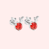 Silver Rudolph earrings with red gemstones on a light pink background.