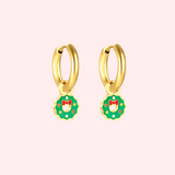 Gold hoop earrings with green wreath charms featuring red bows on a light pink background