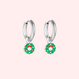Silver hoop earrings with green wreath charms featuring red bows on a light pink background