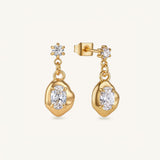 A pair of gold dangle earrings with bezel-set clear marquise-cut gemstones and small floral gold accents, shown on a clean off-white background.