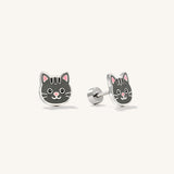 A pair of silver stud earrings shaped like a cute gray cat face, shown on a light cream background. One earring is facing forward to show the detailed cat face with pink inner ears, black stripes, and a pink nose.