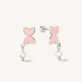 A pair of silver stud earrings featuring a double-heart design with light pink enamel and a dangling white pearl bead, shown on a light cream background.