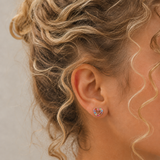 A close-up of a girl's ear wearing a small heart-shaped stud earring with a colorful mosaic pattern on a gold-tone base.