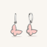 A pair of silver huggie hoop earrings with dangling butterfly charms. The butterfly charms feature a light pink enamel center and a border of small silver beads, set against a plain off-white background.