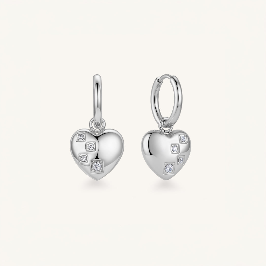A pair of silver heart-shaped hoop earrings with small clear gemstones, shown on a light cream background.