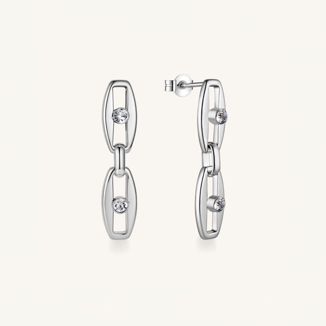 A pair of silver-toned geometric dangle earrings with open oval links and small clear gemstones, shown on a clean off-white background.