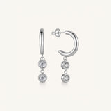 A pair of silver-toned huggie hoop earrings with dangling triple-stone drops, shown on a clean off-white background.