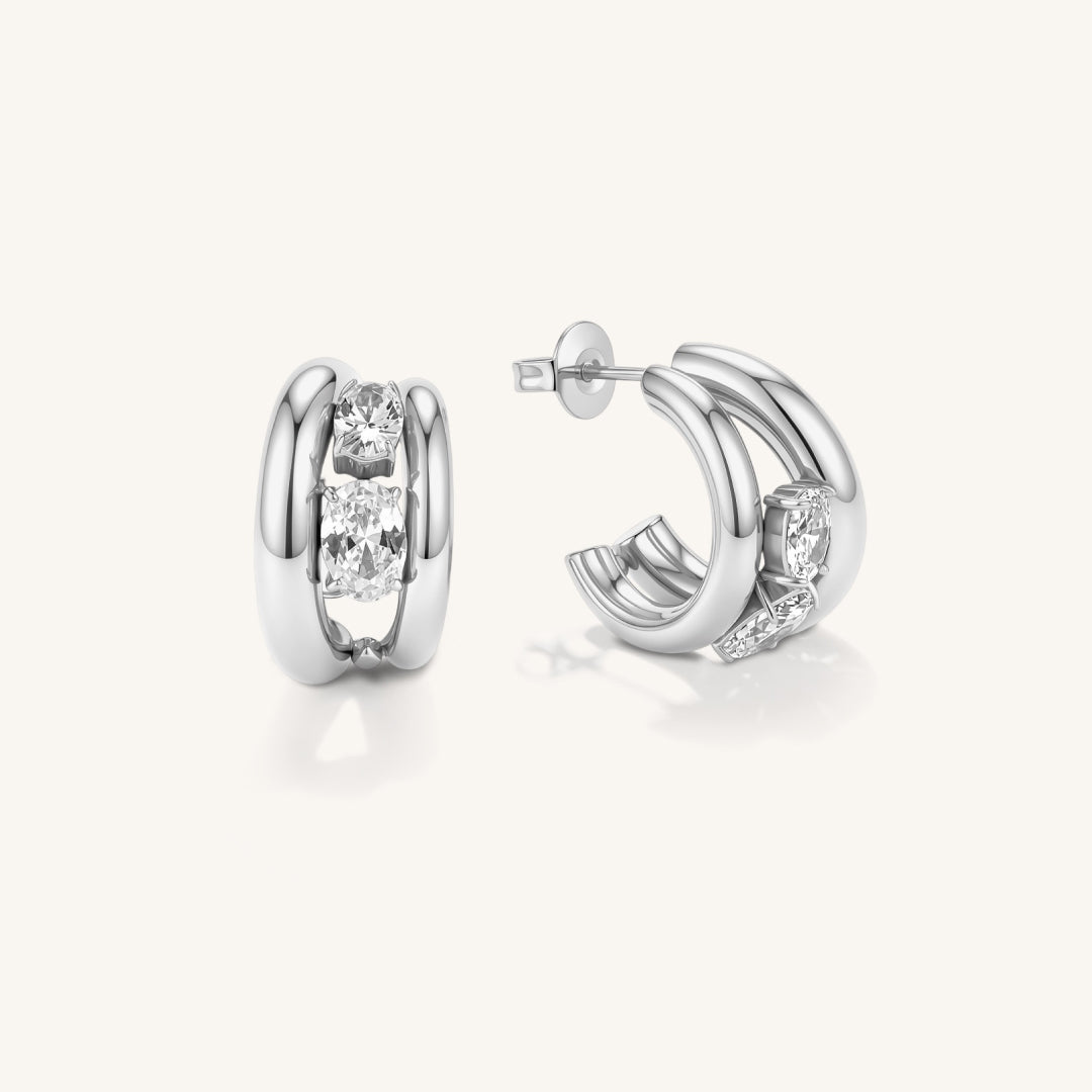 A pair of polished silver hoop earrings with a central channel set with clear round-cut gemstones, shown from both front and side angles on a light cream background.