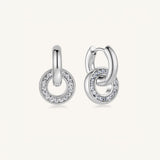 A pair of silver huggie hoop earrings with circular drop elements paved with small clear stones, shown on a light cream background.