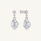 A pair of silver dangle earrings with oval-cut clear gemstones and star-shaped stud accents, shown on a light cream background.