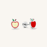 A pair of silver stud earrings shaped like apple slices, one showing the front with red enamel, a pale yellow center, and green leaf accents, and the other showing the side profile with a red apple face and silver post, on a light cream background.