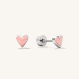 A pair of silver heart stud earrings with a textured light pink center, shown from both front and side angles on a light cream background.