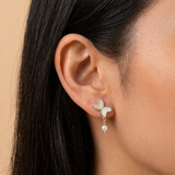 A close-up of a person's ear wearing a small butterfly-shaped stud earring with silver enamel, a pearl drop against a warm beige background.